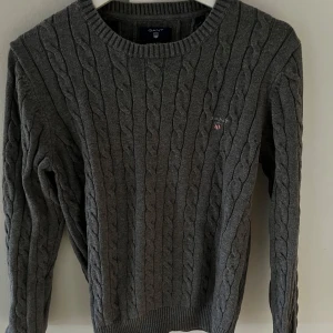 Grå kabelstickad tröja från GANT - Säljer en stilren grå kabelstickad tröja från GANT. Tröjan har en rund halsringning och ett diskret broderat märke på bröstet. Perfekt för kyligare dagar och ger en klassisk look.