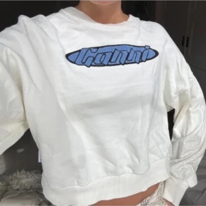 Vit sweatshirt från Ganni - Säljer en vit sweatshirt från Ganni med blått logotryck på bröstet. Tröjan har långa ärmar och en avslappnad passform. Perfekt för en casual look.