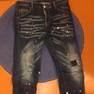 Dsquared2 Jeans - Snygga mörkblå jeans från Dsquared2 med coola slitningar och färgstänk för en unik look. Jeansen har en klassisk femficksdesign och en knappgylf. Perfekta för dig som vill sticka ut med stil. Äkta såklart