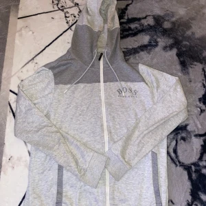 Grå hoodie från Hugo Boss - Snygg grå hoodie från Hugo Boss med dragkedja och huva. Tröjan har ett stilrent märke på bröstet och text längs ärmarna. Perfekt för en avslappnad stil.