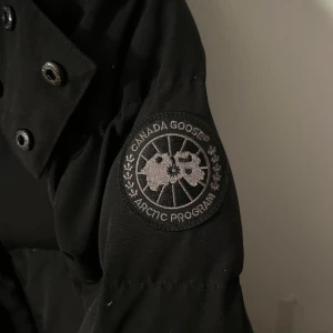 Svart dunjacka från Canada Goose - Säljer en svart dunjacka från Canada Goose med lång ärm och knappar. Jackan har en broderad logga på ärmen och är perfekt för kalla dagar. Den är både stilren och funktionell med en klassisk design. Priset går o diskuteras!!