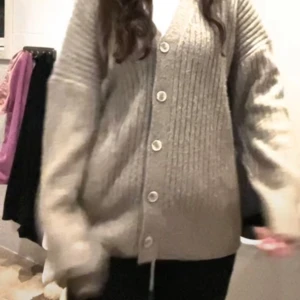 Beige stickad kofta med knappar - Mysig beige stickad kofta med knappar framtill. Koftan har en ribbad struktur och långa ärmar, perfekt för kyligare dagar. Den har en avslappnad passform som ger en bekväm känsla.