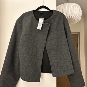 Grå jacka/kappa - Snygg grå jacka från Gina tricot i en stilren design. Jackan har en enkel knäppning framtill och en rund halsringning. Perfekt för en chic och minimalistisk look.