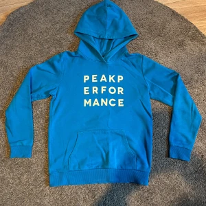 Blå hoodie från Peak Performance - Säljer en snygg blå hoodie från Peak Performance med text på framsidan. Tröjan har en klassisk känguruficka och en bekväm luva. 