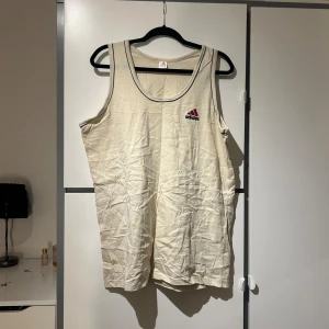 Beige linnetopp från Adidas - Säljer en beige linnetopp från Adidas med klassisk logga i rött och svart på bröstet. Toppen har en enkel och stilren design med svarta kantdetaljer.
