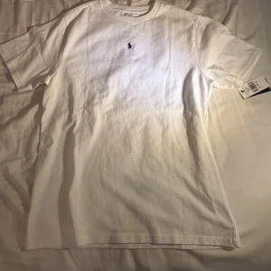 Vit t-shirt från Ralph Lauren - Säljer en klassisk vit t-shirt från Ralph Lauren med det ikoniska broderade logotypen på bröstet. T-shirten är kortärmad och tillverkad i mjuk bomull för en bekväm passform. Perfekt för en stilren och avslappnad look. Pris kan diskuteras. Den är äkta såklart. Köpt på kidsbrandstore. Köpt för 595kr