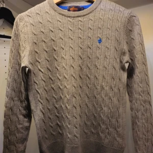 Kabelstikad Morris tröja  - Säljer en stilren beige stickad tröja med kabelstickat mönster och en liten blå broderad logga på bröstet. Tröjan har långa ärmar och rund halsringning. Perfekt för kyligare dagar! Bra skick, priset går att förhandla.