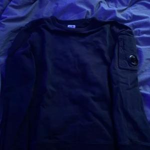 Svart tröja från C.P. Company - Säljer en stilren marinblå tröja från C.P. Company med en unik detalj på ärmen. Tröjan har långa ärmar och en rund halsringning. Perfekt för en casual look. Nypris 2000kr