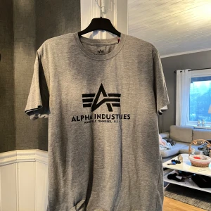Grå t-shirt från Alpha Industries - Säljer en grå t-shirt från Alpha Industries med deras ikoniska logga tryckt på framsidan. T-shirten är kortärmad och tillverkad i mjukt material, perfekt för en avslappnad stil.