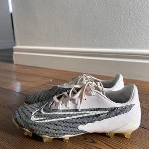 Grå och vita fotbollsskor från Nike - Snygga fotbollsskor från Nike i grått och vitt med snörning. Skorna har en modern design med ett randigt mönster och är perfekta för fotbollsplanen. De har en klassisk Nike-logga på sidan.