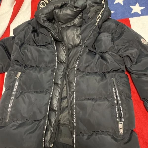 Svart dunjacka från Moncler - Säljer en svart dunjacka från Moncler med broderad logga på ärmen. Jackan har en hög krage och dragkedja framtill. Perfekt för kyliga dagar med sin pufferdesign och stilrena look.