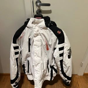  Vinterjacka Geographical Norway Strl L - Säljer en cool vit och svart vinterjacka från Geographical Norway. Jackan har en huva och flera praktiska fickor med dragkedjor. Den har snygga röda detaljer och är perfekt för kalla dagar. Jackan är designad för att hålla dig varm och bekväm under vintern.  Obs jackan är aldrig använd!