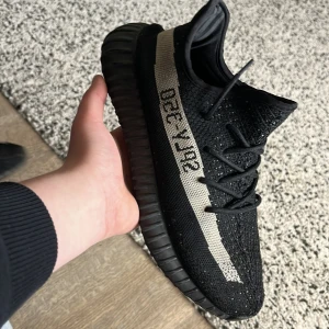 Svarta Adidas Yeezy sneakers - Snygga svarta Adidas Yeezy sneakers med en distinkt beige rand och text på sidan. Skorna har en robust sula och är designade för både stil och komfort. Perfekta för dig som vill ha en trendig look.