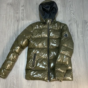 Grön pufferjacka från Moncler - Säljer en snygg grön pufferjacka från Moncler med glansig yta och huva med pälsdetaljer. Jackan har dragkedja framtill och två fickor med dragkedjor. Perfekt för kyliga dagar.