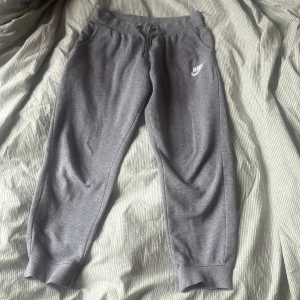Grå mjukisbyxor från Nike . - Säljer ett par grå mjukisbyxor från Nike med elastisk midja och snörning. Byxorna har ribbade muddar vid bensluten och en klassisk Nike-logga på benet. Perfekta för en avslappnad stil.