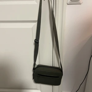 Olivgrön axelväska med dragkedja - Snygg olivgrön axelväska med justerbar rem och praktisk dragkedja framtill. Perfekt för att bära dina nödvändigheter med stil. Enkel och stilren design som passar till många tillfällen.