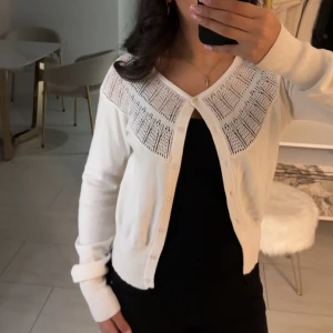 Vit kofta med spetsdetaljer - Säljer en elegant vit kofta med spetsdetaljer vid axlarna. Koftan har långa ärmar och knappar framtill. Perfekt för en stilren look. den här en fläck vid armen som jag inte riktigt vet hur den uppstått jag vet inte heller vilket märke det är då jag klippt av lappen men om jag minns rätt så var det antingen h&m eller lindex