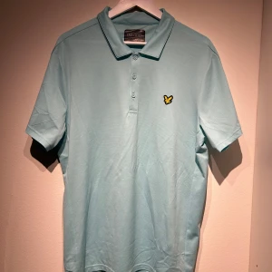 Ljusblå pikétröja från Lyle & Scott - Snygg ljusblå pikétröja från Lyle & Scott med klassisk krage och knappslå. Tröjan har ett broderat gult emblem på bröstet och korta ärmar. Perfekt för en stilren och avslappnad look.