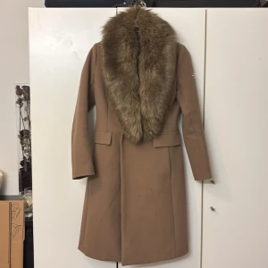 Beige kappa med pälsdetalj från Hollies - Elegant beige kappa från Hollies med en lyxig pälsdetalj runt kragen. Kappan har en klassisk design med knappar framtill och fickor på sidorna. Perfekt för att hålla stilen under kyliga dagar.