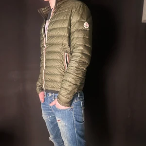 Grön dunjacka från Moncler - Säljer en snygg grön dunjacka från Moncler.. Perfekt för både kyliga och lite varmare dagar med sin stilrena look och bekväma passform. Jackan är i okej skick den har lite lagningar som är med på bilderna och tyvärr även lite mer små hål alltså inte perfekt skick där av priset!
