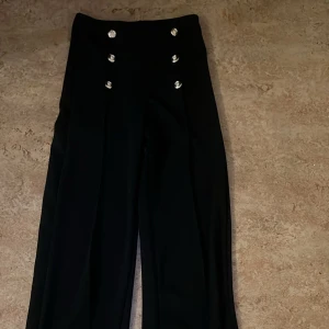 Chiquelle byxor med knappar - Snygga svarta byxor med hög midja och dekorativa knappar framtill. Byxorna har en elegant och stilren design med vida ben som ger en chic look. Perfekta för att lyfta vilken outfit som helst.