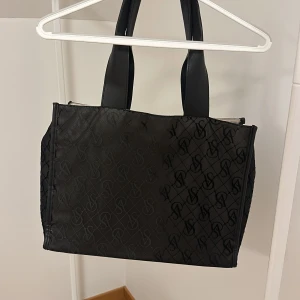 Svart shoppingväska med monogram - Snygg svart shoppingväska med monogrammönster. Väskan har två rejäla handtag och är perfekt för att bära med sig allt man behöver. Stilren och praktisk design som passar till många tillfällen.