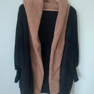 Mysig teddyjacka i svart och beige - Säljer en supermysig teddyjacka i svart med beige insida. Perfekt för kyliga dagar när du vill känna dig som en vandrande kram. Jackan har långa ärmar och en lös passform som gör den extra bekväm. Den är i mycket bra skick och redo för nya äventyr!