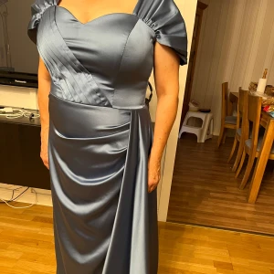 Satinklänning - Vacker Blå/grå klänning i satin. Endast använd en gång. Köpt på Sasha i Södertälje. Nypris 2600kr. Storlek M, personen på bilden är 159cm. Den har en slit vid benet som täcks av släpet.