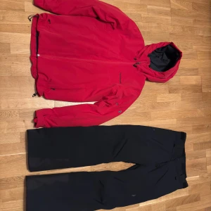 Röd och svart skidställ från Peak Performance - Säljer en snygg röd och svart skidställ från Peak Performance. Perfekt för skidåkning men även för vardaglig användning i kalla vinterdagar med sin tåliga polyester och praktiska dragkedja. Jackan har en justerbar huva och långa ärmar, vilket gör den idealisk för utomhusaktiviteter.                                                         Storlek S på byxorna och M i jackan.                                      Ser exakt ut som ny då den endast användes för en vecka för skidåkning i Sälen.