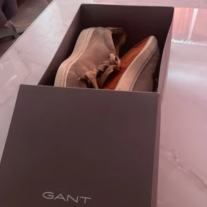 Beige sneakers från GANT - Säljer ett par snygga beige sneakers från GANT. De är i mocka med en vit sula och har snörning. Perfekta för en avslappnad stil och passar bra till både vår och höst. Skorna är i bra skick och redo för nya äventyr!