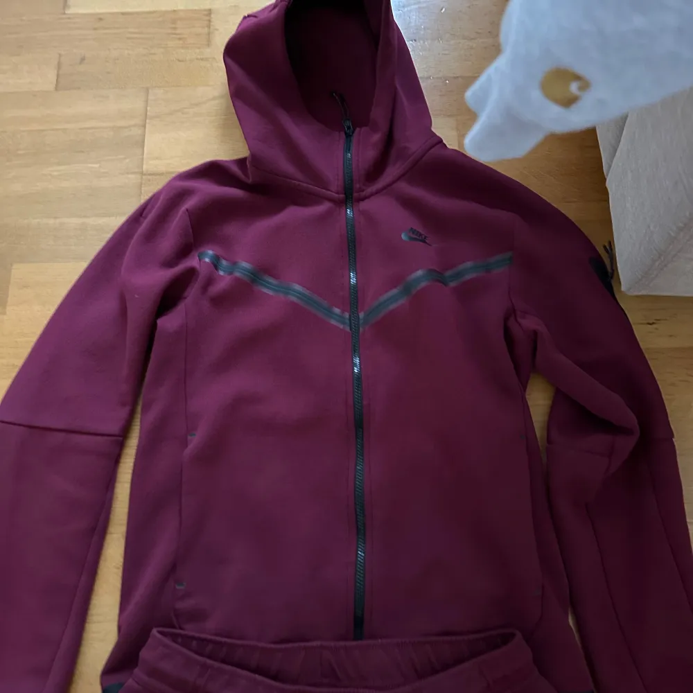 Säljer ett set med vinröd hoodie och matchande byxor från Nike. Hoodien har dragkedja och en snygg detalj i form av ett grått mönster över bröstet. Byxorna har en bekväm passform med dragsko i midjan. Perfekt för en avslappnad stil eller träning. Storlek S i koftan och M i byxorna.. Tröjor & Koftor.