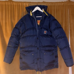 Blå dunjacka från Fjällräven - Säljer en snygg blå dunjacka från Fjällräven i storlek S. Jackan är perfekt för kalla vinterdagar och har en praktisk huva. Den är i mycket bra skick och har både dragkedja och knappar framtill. Till köper medföljer även fjällräven bag och extra knappar.