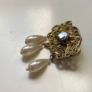 Vintage brosch med pärlor och  sten - Säljer en elegant vintage brosch i guldmetall med tre hängande pärlor och en sten i mitten. Perfekt för att ge en klassisk touch till din outfit. Broschen har en intrikat design och är i fint skick.