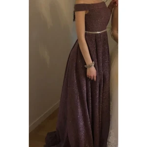 Glittrig festklänning - Säljer en fantastisk glittrig  långklänning med offshoulder-design. Klänningen har en elegant silhuett med en glittrande detalj i midjan som ger en extra touch av glamour. Perfekt för festliga tillfällen som nyår eller bal. Har endast använt en gång. Pris kan disskuteras! 