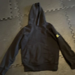 Svart hoodie från Stone Island Junior - Säljer en svart hoodie från Stone Island Junior. Den har en klassisk design med en stor ficka fram och en huva. Märkets ikoniska logga sitter på ärmen. Perfekt för en stilren och bekväm look. Passar både till vardags och chill.