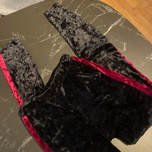 Svarta sammetbyxor med röda detaljer - Snygga svarta byxor i sammet med röda detaljer längs sidorna. Perfekta för en festlig kväll eller en mysig vinterdag. Byxorna har en elegant look och känns supermjuka. Passar perfekt för dig som vill ha något unikt och stilrent.
