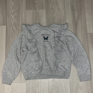 Grå tröja med volanger från H&M - Säljer en superfin grå tröja från H&M med texten 'Ready to Fly' och en söt fjäril på framsidan. Tröjan har långa ärmar och volangdetaljer som ger den en extra touch.Perfekt men tyvärr passar inte längre.Materialet är mjukt och bekvämt, perfekt för höst och vår!💕☺️