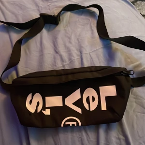 Svart axelväska från Levi's - Säljer en snygg svart axelväska/magväska från Levi's. Väskan har en justerbar rem och ett stort vitt tryck med Levi's logga på framsidan. Perfekt för vardagsbruk och rymmer det mesta du behöver ha med dig. Den har en dragkedja för säker förvaring.