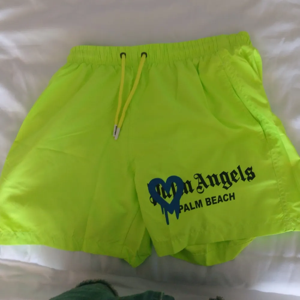 Säljer ett par supercoola neongula shorts från Palm Angels med en blå hjärtdetalj och texten 'Palm Beach'. De har en elastisk midja med gul dragsko för perfekt passform. Perfekta för sommardagar på stranden eller bara chilla runt i stan. 🏖️. Shorts.