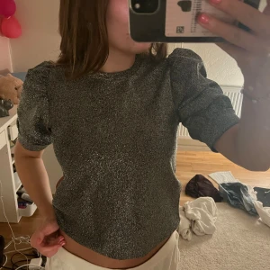 Glittrig topp med puffärmar - Säljer en superfin glittrig topp i silver med puffärmar. Den är kortärmad och, perfekt för festliga tillfällen. Toppen är i nyskick och redo att ge lite extra bling till din outfit!