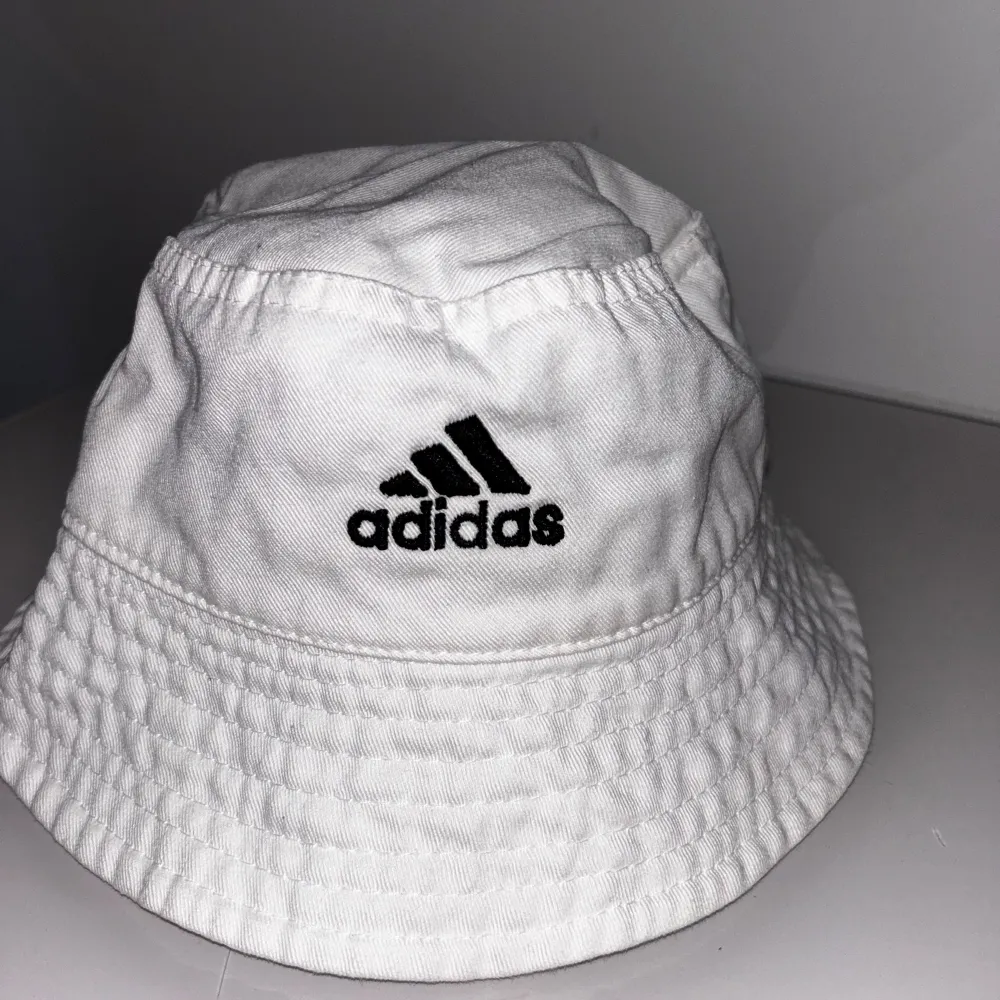 Säljer en snygg vit bucket hat från Adidas. Den har en klassisk stil med Adidas-loggan broderad i svart framtill. Perfekt för sommardagar eller som en cool accessoar till din outfit.. Accessoarer.