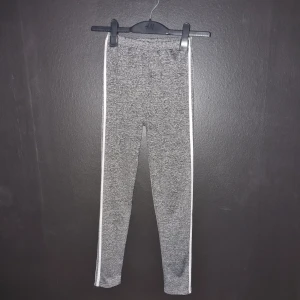 Grå leggings med vita ränder - Säljer ett par grå träningsleggings med vita ränder längs sidorna. De är superbekväma och perfekta för träning. Materialet är stretchigt och mjukt, vilket gör dem sköna att ha på sig hela dagen.