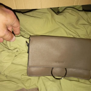 Beige axelväska från NYPD - Säljer en snygg beige axelväska från NYPD. Väskan är i skinn och har en stilren design med en stor ringdetalj på framsidan. Insidan är mönstrad och har en praktisk dragkedja. Perfekt för både vardag och fest!