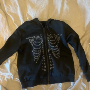 Svart hoodie med skelettmönster - Säljer en svart hoodie med dragkedja och ett coolt skelettmönster i silverfärgade nitar på framsidan. Perfekt för höst och vinter, och den är långärmad för extra värme. Passar bra till vardags eller fest!