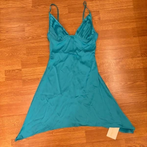 Turkos klänning med smala axelband - Säljer en snygg turkos klänning i satin med smala axelband. Klänningen har en asymmetrisk nederkant och en v-ringad design som ger en elegant look. Perfekt för sommarens fester eller en kväll ute! För stor för mig och därav oanvänd.