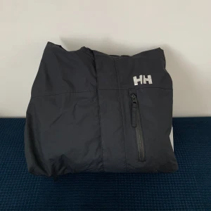 Svart jacka från Helly Hansen - Säljer en svart jacka från Helly Hansen i storlek S. Jackan har en dragkedja och en praktisk huva med dragsko. Perfekt för kyliga dagar och har en stilren design med Helly Hansen-loggan på bröstet. Nypris 1599kr. 