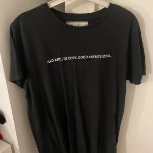 säljer en svart t-shirt med texten 'BAD ARTISTS COPY. GOOD ARTISTS STEAL.' på framsidan. på baksidan finns ett svartvitt porträtt av en man. t-shirten är kortärmad och i normal passform. unisex