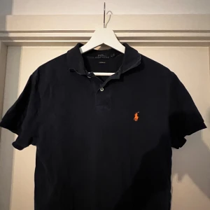 Svart pikétröja från Ralph Lauren - Säljer en klassisk svart pikétröja från Ralph Lauren i storlek M. Tröjan har en snygg orange logga på bröstet och är i custom fit. Perfekt för både vardag och fest!