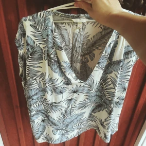 Mönstrad blus från H&M - Säljer en snygg blus från H&M med ett tropiskt bladmönster i blått och vitt. Blusen har en v-ringad design och korta ärmar, perfekt för sommardagar. Materialet känns lätt och luftigt, vilket gör den idealisk för varma väder.