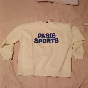 Säljer en vit sweatshirt från H&M Divided med texten 'PARIS SPORTS' i blått på framsidan. Tröjan är i storlek M och har långa ärmar. Perfekt för en avslappnad stil eller sportiga dagar. Den är i bra skick och redo för nya äventyr!